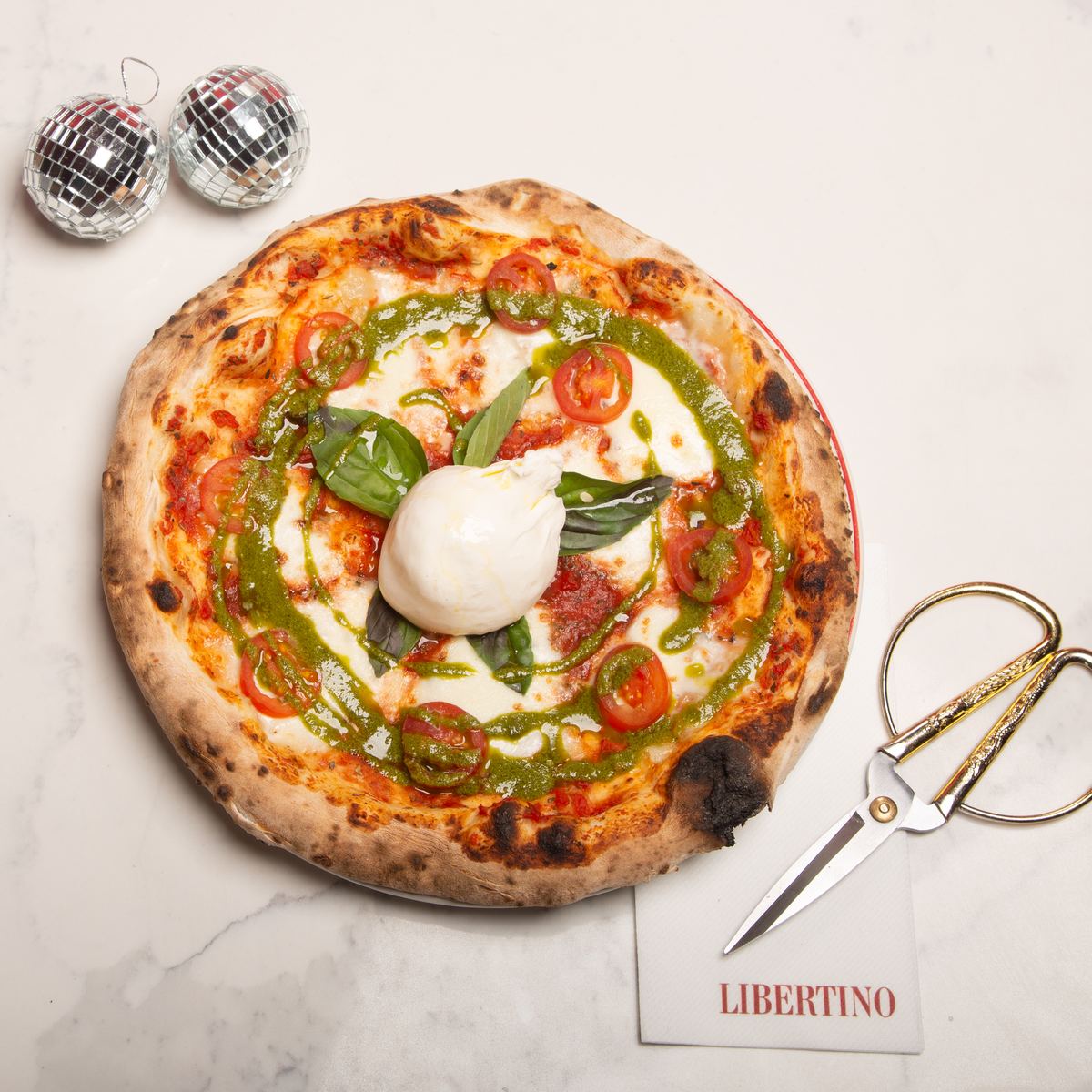 A Neapolitan pizza with burrata, pesto and basil, gold scissors and two small disco balls beside it